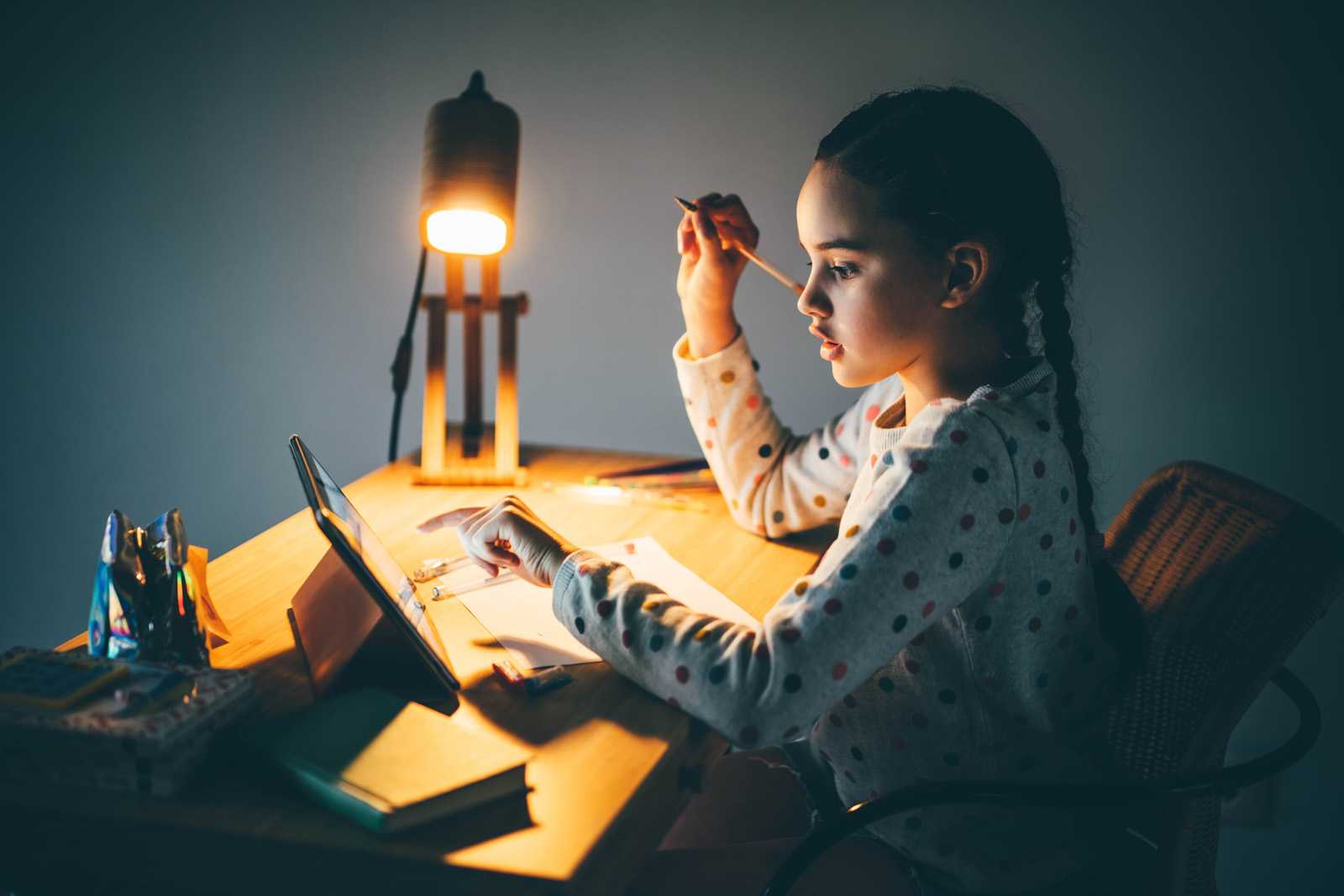 girl sitting at desk at night with lamp on, completing lesson on a tablet and work book