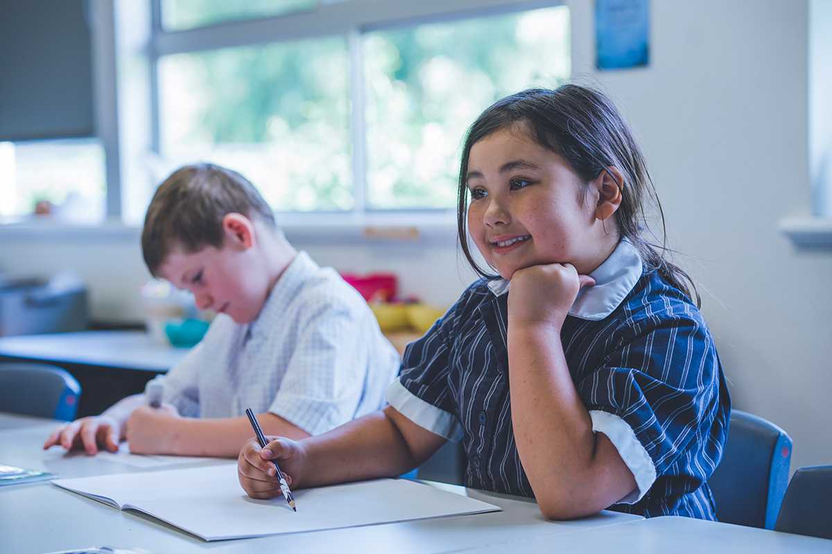 Smiling primary aged girl sitting at desk with head resting on hand